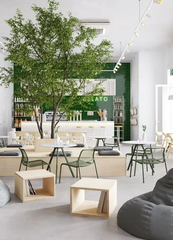 Modern cafe interior with minimalist furniture, green accents, and tree centerpiece, creating a cozy atmosphere.
