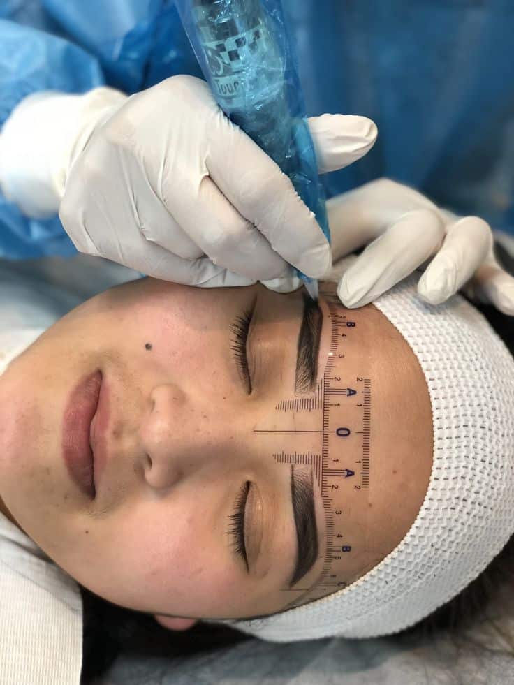 _ 1 Person undergoing precise eyebrow tattooing with a blue pen and ruler guide, wearing a white headband. | Sky Rye Design Person undergoing precise eyebrow tattooing with a blue pen and ruler guide, wearing a white headband.