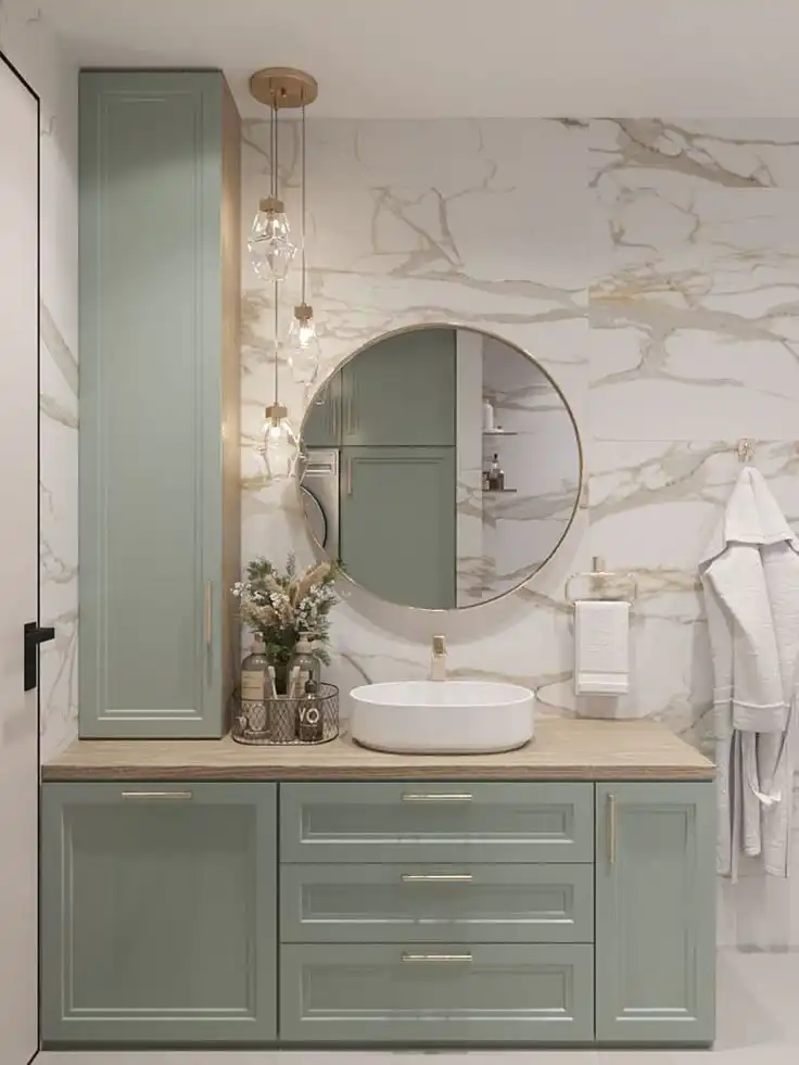 Modern bathroom with green cabinets, round mirror, and marble walls. Features pendant lighting and a countertop vessel sink