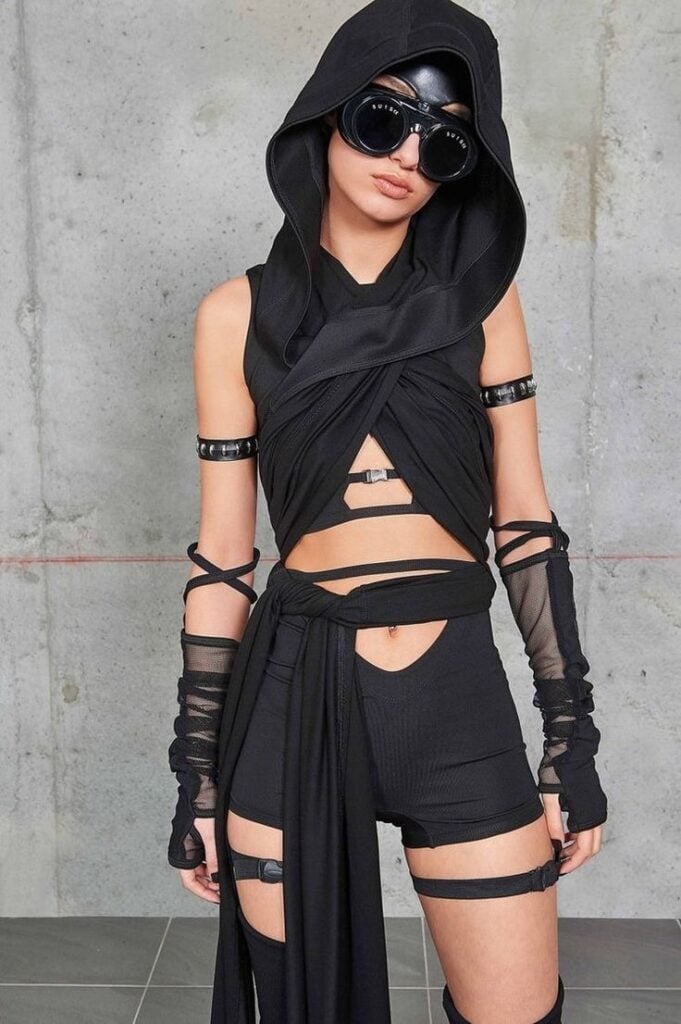 _ 15 Fashionable individual in futuristic black outfit with hood, sunglasses, and arm wraps against a concrete wall backdrop. | Sky Rye Design Fashionable individual in futuristic black outfit with hood, sunglasses, and arm wraps against a concrete wall backdrop.