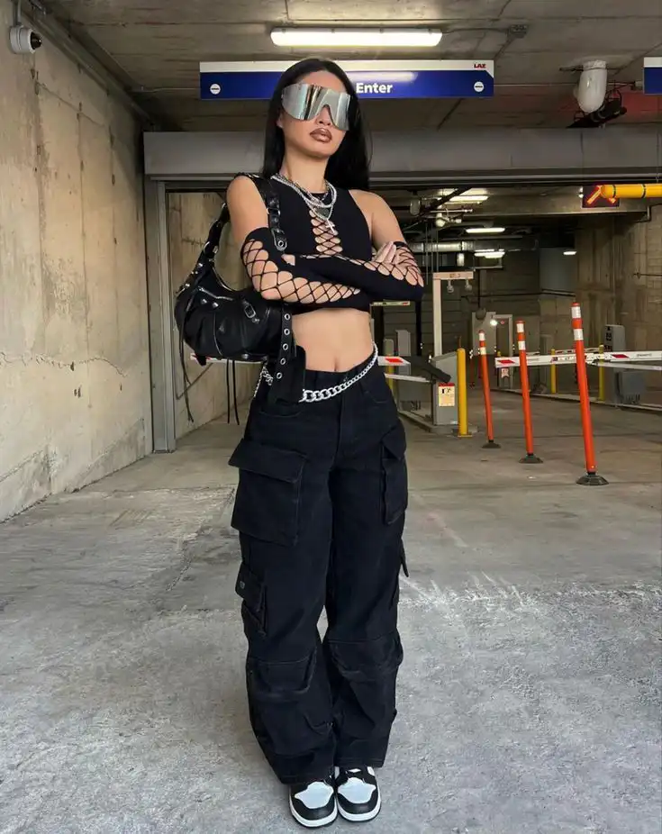 Trendy woman with futuristic sunglasses, black cargo pants, and fishnet top, posing confidently in urban setting.