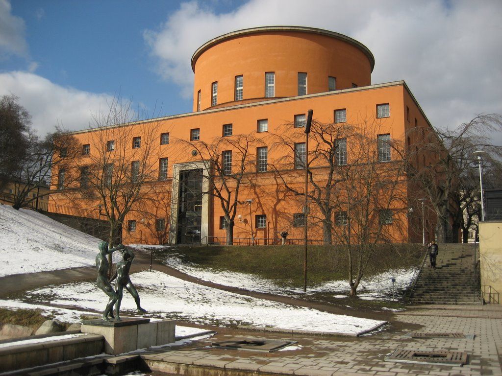 Tall, round, orange building with trees and statue in foreground, under cloudy sky, partially snowy ground.