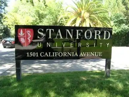 Stanford University entrance sign at 1501 California Avenue, surrounded by greenery and a palm tree in the background on a sunny day.
