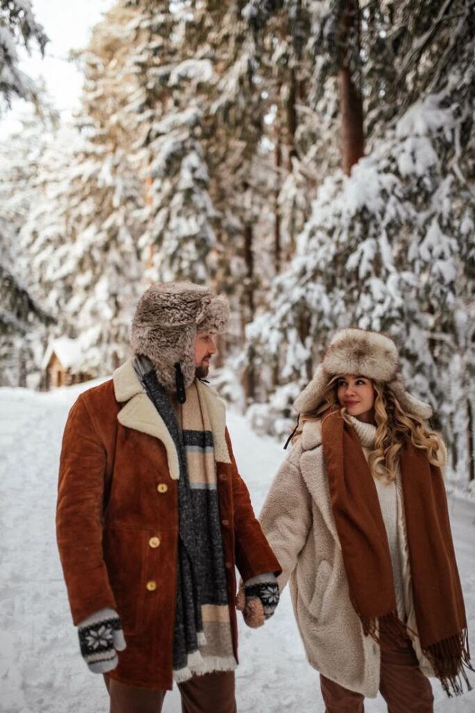 _ 2 Couple walking hand in hand in snowy forest, dressed warmly in brown coats and wool hats, showcasing winter fashion amidst snowy trees. | Sky Rye Design Couple walking hand in hand in snowy forest, dressed warmly in brown coats and wool hats, showcasing winter fashion amidst snowy trees.