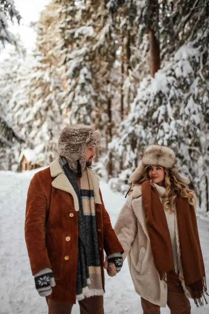 _ 2 Couple walking hand in hand in snowy forest, dressed warmly in brown coats and wool hats, showcasing winter fashion amidst snowy trees. | Sky Rye Design Couple walking hand in hand in snowy forest, dressed warmly in brown coats and wool hats, showcasing winter fashion amidst snowy trees.