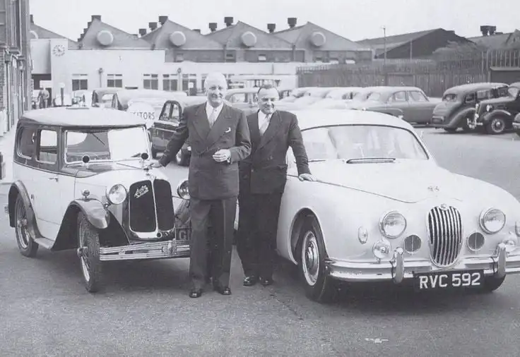 Two men in suits stand proudly between a vintage and a classic car, highlighting automotive history and evolution in a