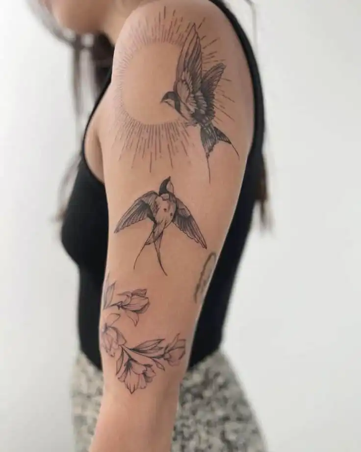 Female arm showcasing a black bird and flower tattoo design, featuring sunburst detail, against a plain white background.