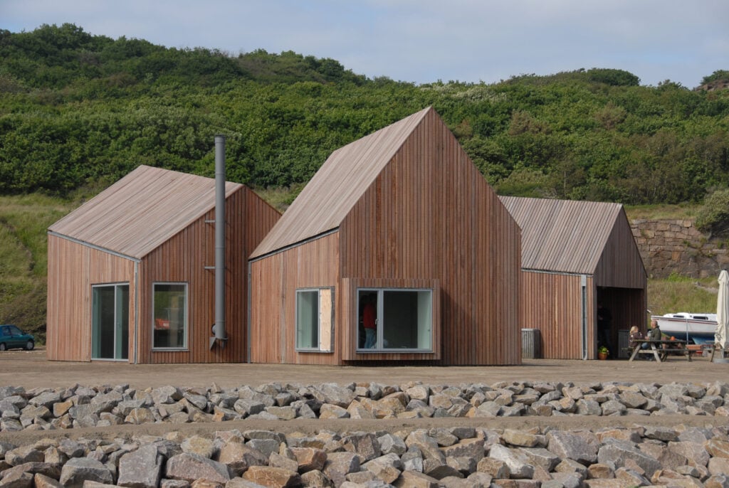 Modern wooden cabins with sloped roofs, surrounded by lush greenery and rocks, under a clear sky.