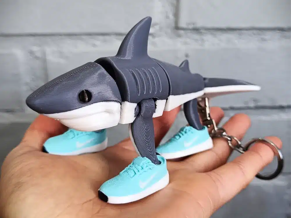 3D-printed shark keychain with teal sneakers on a hand, against a gray brick wall backdrop. Unique and quirky accessory.