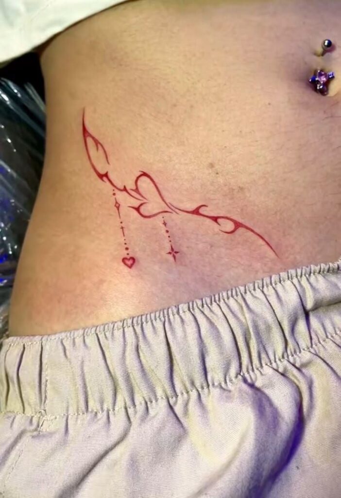 _ - 2026-01-04T191013266 Close-up of a red ink decorative tattoo on the side of a person's torso, featuring abstract shapes and hanging heart details. | Sky Rye Design Close-up of a red ink decorative tattoo on the side of a person's torso, featuring abstract shapes and hanging heart details.