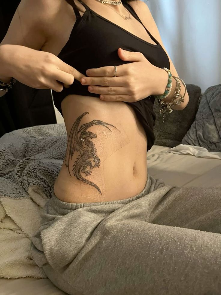 _ - 2026-01-04T191612121 Person showing a detailed dragon tattoo on their side, wearing a black top and grey pants, sitting on a bed with cozy blankets. | Sky Rye Design Person showing a detailed dragon tattoo on their side, wearing a black top and grey pants, sitting on a bed with cozy blankets.
