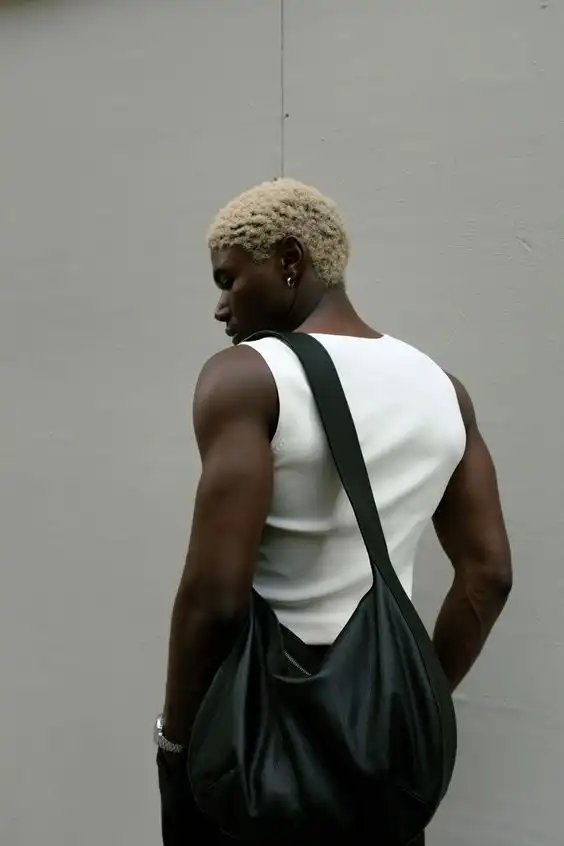 Stylish person with short hair and a sleeveless top carrying a black bag, standing against a neutral background. Fashion-forward look.