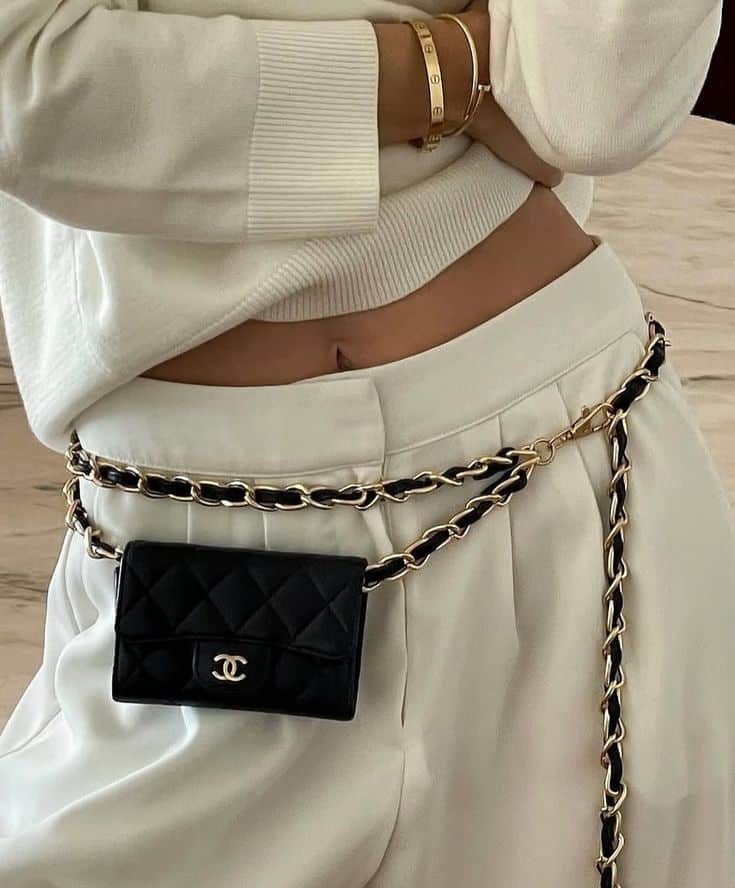 Woman in white outfit accessorized with a black quilted mini bag and gold chain belt, showcasing a chic and stylish fashion look.