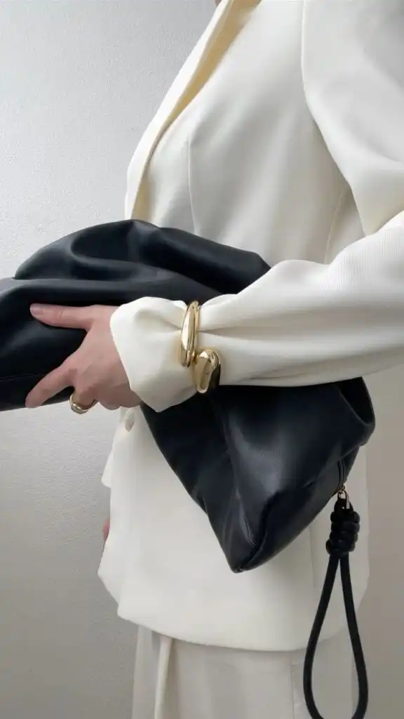 Person in a stylish white suit holding a black leather clutch, accessorized with gold bangles. Elegant, modern fashion.