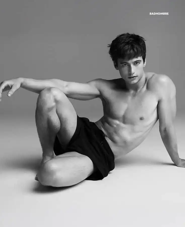 Shirtless man seated on the floor in black shorts, posing confidently in a black-and-white studio setting.