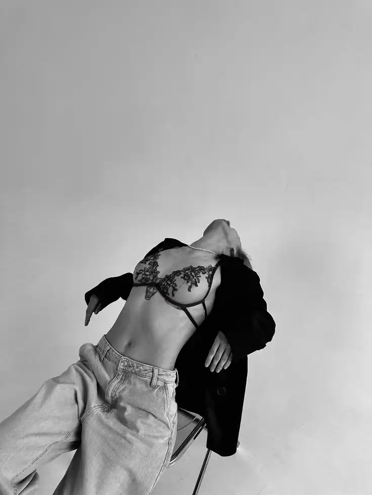 Stylish woman in lace lingerie and open jacket poses artistically on a chair in a minimalist black and white setting.