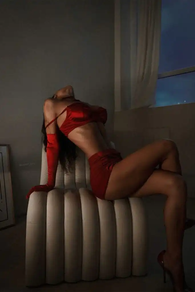 Woman in red lingerie poses dramatically on a stylish chair in a dimly lit room with a window view.