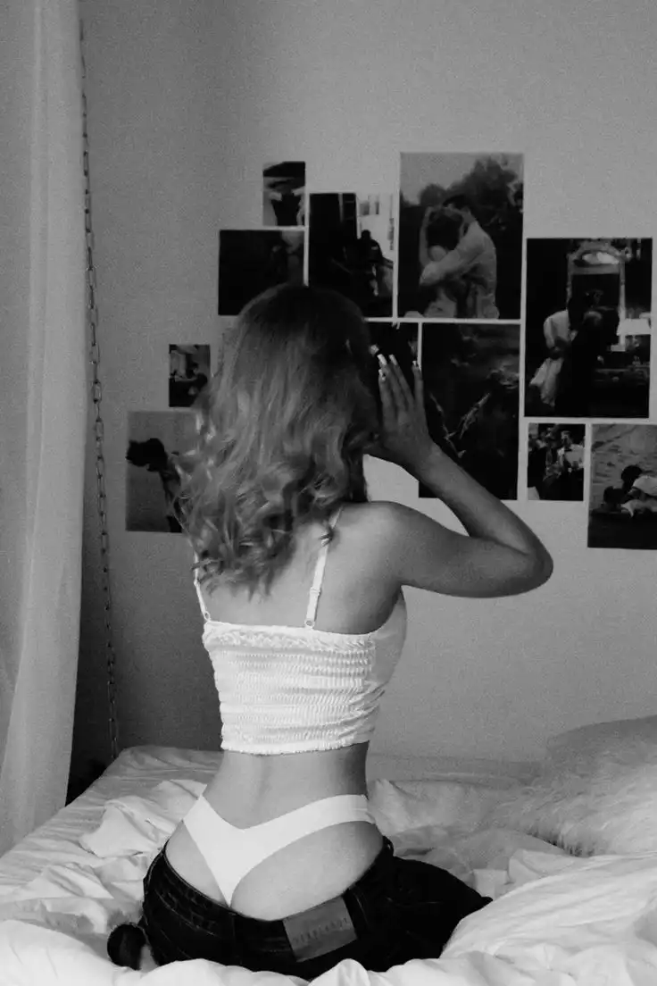 Woman sitting on bed in front of a photo wall, wearing a white top and jeans, with her hand near her face, in black and white.