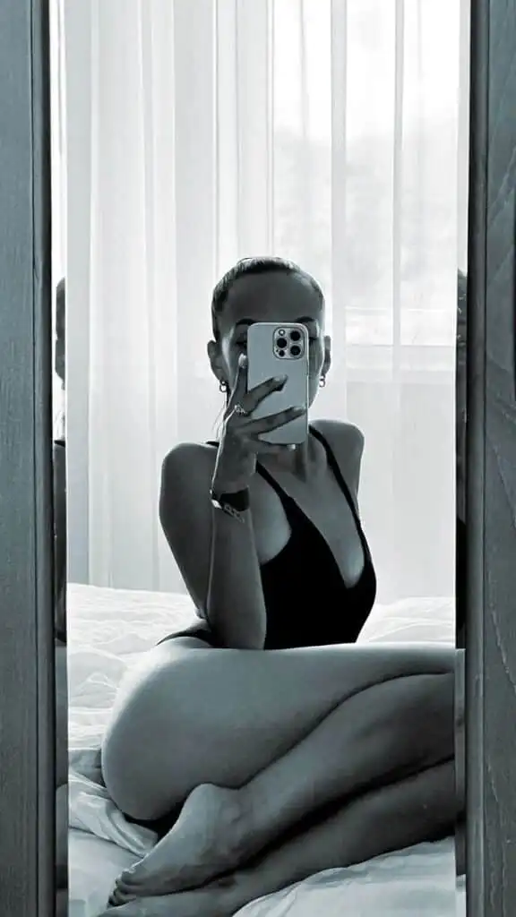 Person in a black swimsuit takes a mirror selfie on a bed, framed by soft, sheer curtains in a minimalist room setting.