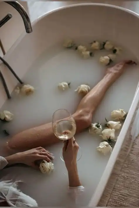Person relaxing in a milky bath surrounded by white roses, holding a glass of wine, creating a serene spa-like atmosphere.