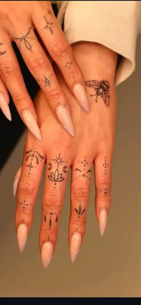 Close-up of hands with intricate finger tattoos and long nude-colored nails, showcasing delicate symbols like stars, moons, and a bee design.