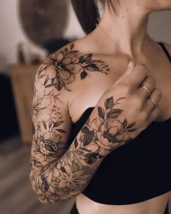Floral tattoo sleeve on a woman's arm and shoulder, featuring intricate flower designs, captured indoors with soft lighting.