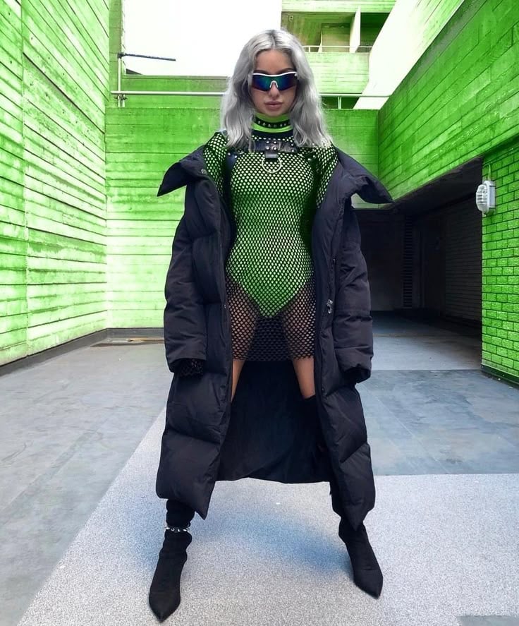 _ 23 Fashion-forward individual in bold green mesh outfit, black puffer coat, and sunglasses, standing in vibrant green urban | Sky Rye Design Fashion-forward individual in bold green mesh outfit, black puffer coat, and sunglasses, standing in vibrant green urban