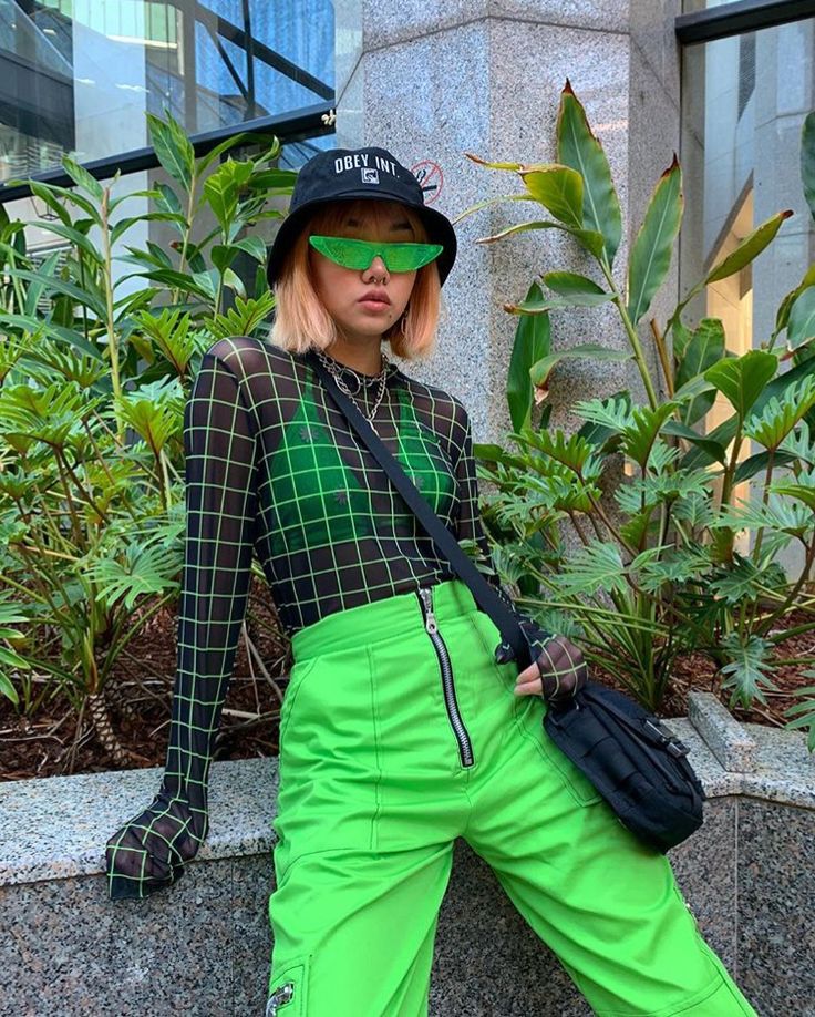 _ 24 Stylish person with green checkered top, neon green pants, sunglasses, and bucket hat in urban setting with lush plants. | Sky Rye Design Stylish person with green checkered top, neon green pants, sunglasses, and bucket hat in urban setting with lush plants.