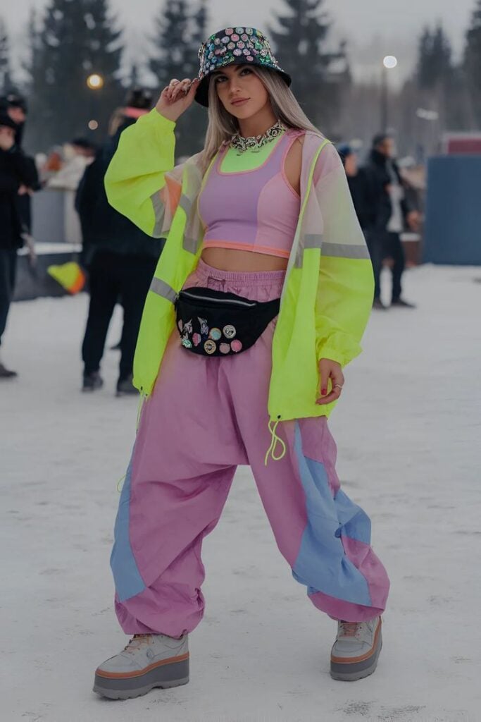 24 Womens Fashionable Winter Rave Outfits For Festivals Young woman in vibrant retro outfit, neon jacket, and stylish bucket hat poses confidently outdoors in a snowy urban setting. | Sky Rye Design Young woman in vibrant retro outfit, neon jacket, and stylish bucket hat poses confidently outdoors in a snowy urban setting.