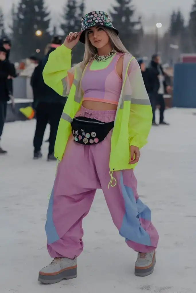 Young woman in vibrant retro outfit, neon jacket, and stylish bucket hat poses confidently outdoors in a snowy urban setting.