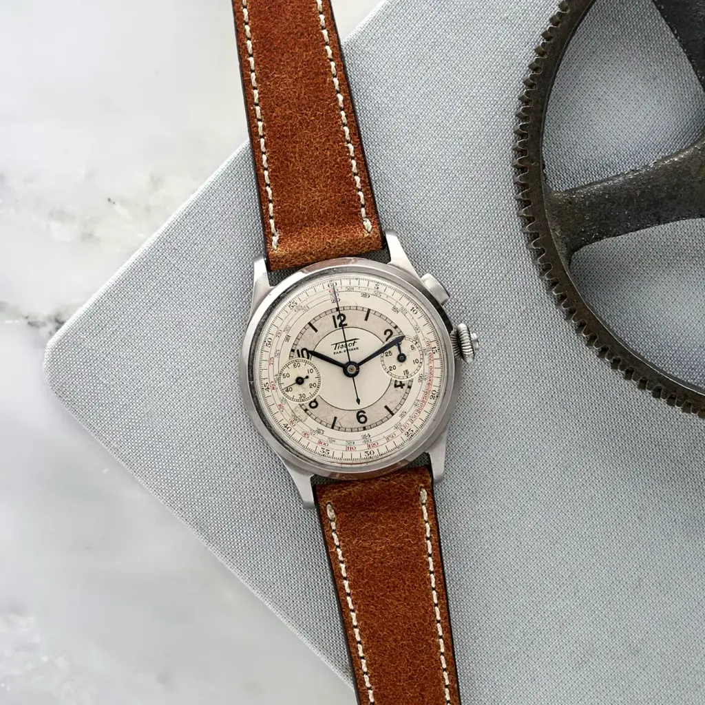 Vintage wristwatch with a brown leather strap on a gray surface next to a gear, showcasing a classic and elegant design.