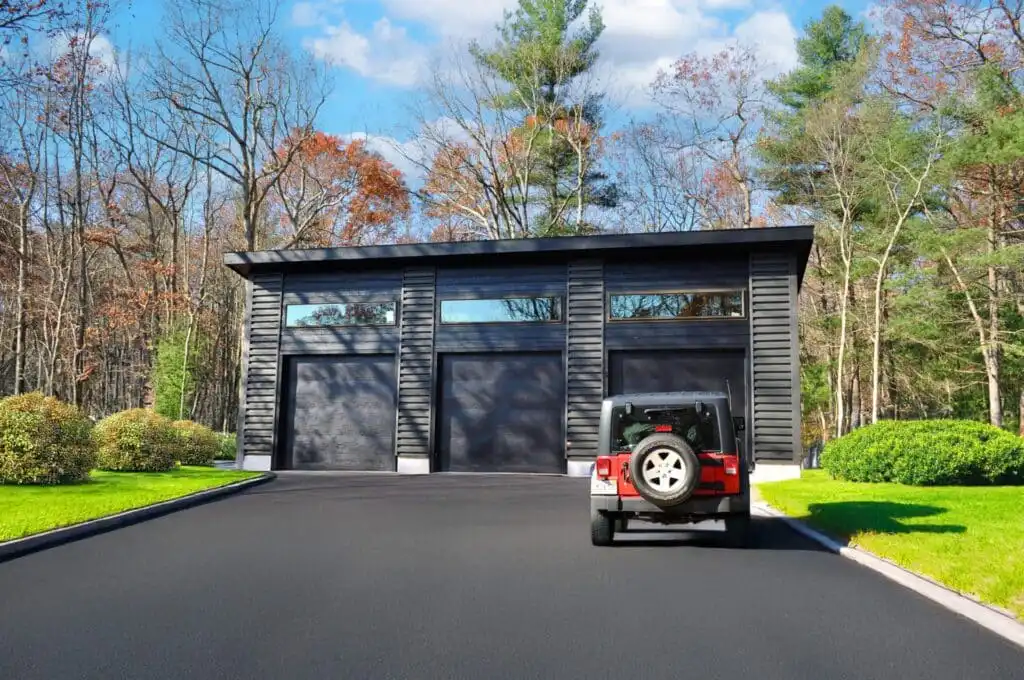 Black modern garage with three doors, surrounded by green trees and shrubs, with a red jeep parked on the driveway.