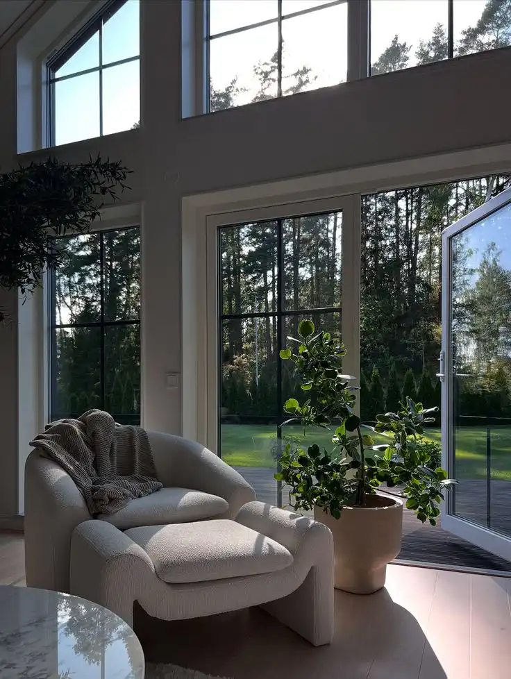 _ 28 Cozy modern interior with armchair, throw blanket, and potted plant by sunlight-filled floor-to-ceiling windows, overlooking a lush garden. | Sky Rye Design Cozy modern interior with armchair, throw blanket, and potted plant by sunlight-filled floor-to-ceiling windows, overlooking a lush garden.