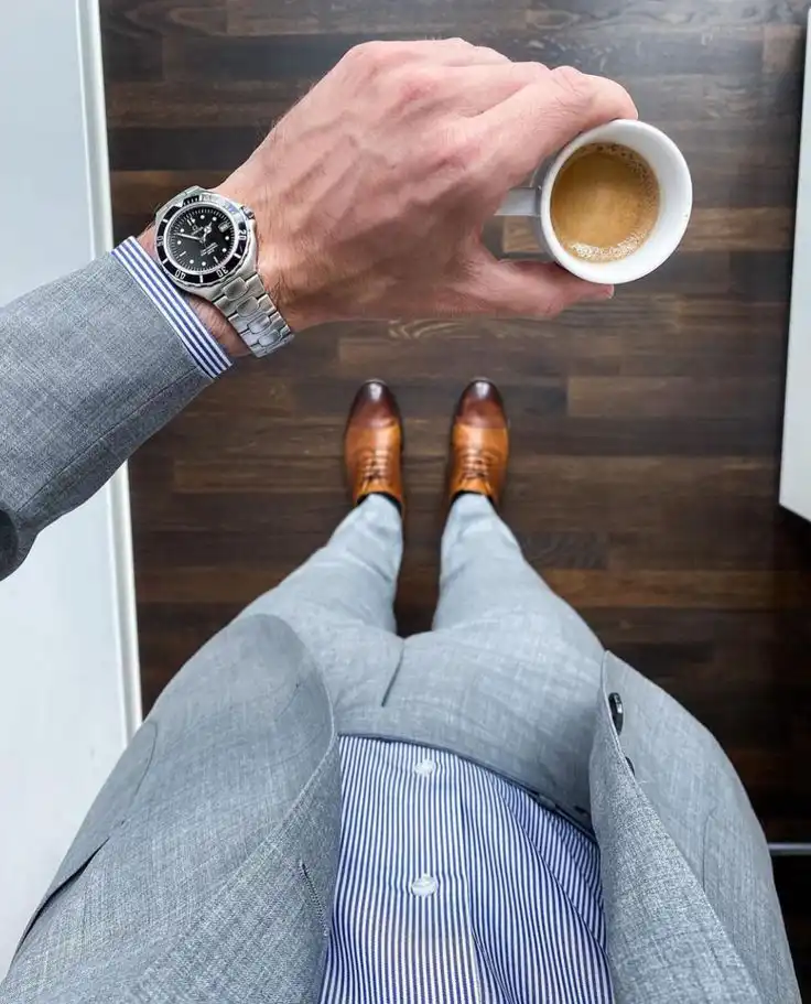 Man in a gray suit holding coffee, wearing a stylish watch and brown shoes on a wooden floor. View from above.