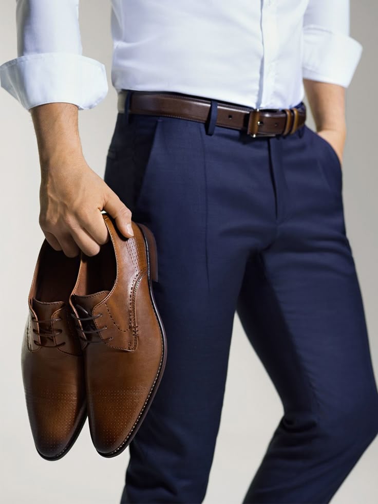 Man in white shirt and navy blue pants holding brown leather dress shoes, showcasing formal fashion style and accessories.