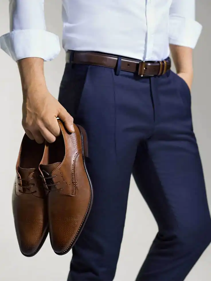 Man in white shirt and navy blue pants holding brown leather dress shoes, showcasing formal fashion style and accessories.