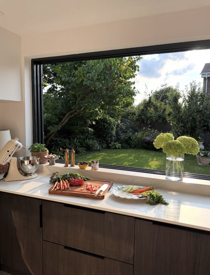 Screenshot Spacious kitchen with a scenic garden view, featuring fresh cut vegetables on a counter and a mixer, bathed in sunlight. | Sky Rye Design Spacious kitchen with a scenic garden view, featuring fresh cut vegetables on a counter and a mixer, bathed in sunlight.