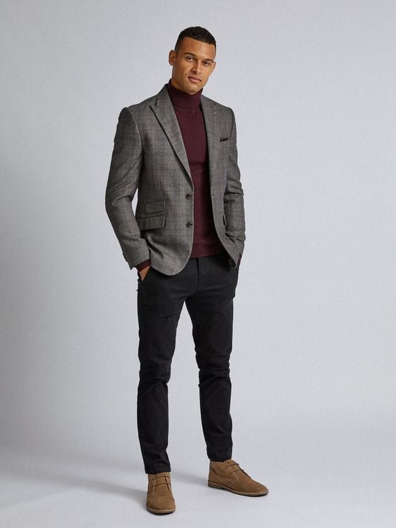 Man in stylish gray blazer and burgundy turtleneck with black pants and brown shoes stands confidently against a neutral