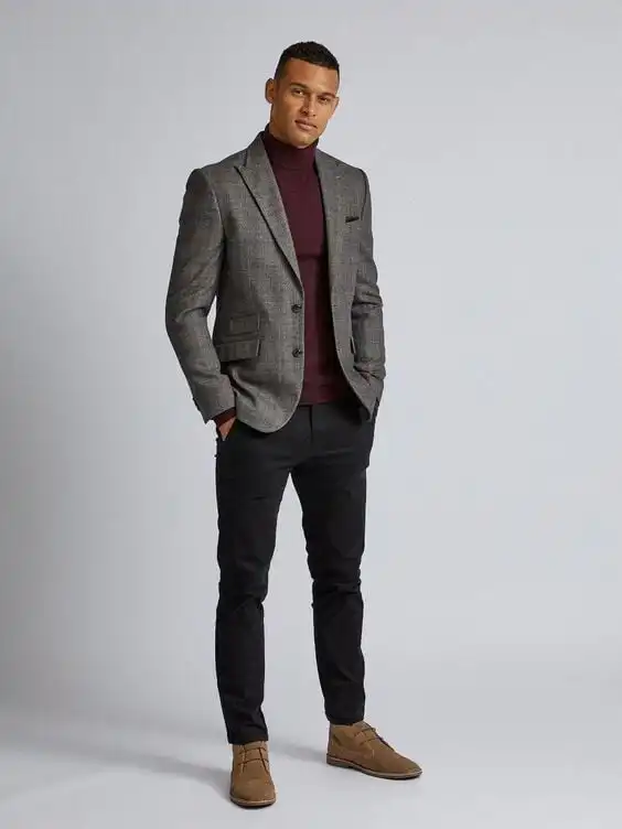 Man in stylish gray blazer and burgundy turtleneck with black pants and brown shoes stands confidently against a neutral