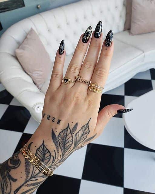 33cdfca1-f70c-4351-915b-65193ae39a34 Tattooed hand with black nail art and gold rings displayed on a sofa backdrop. Elegant gold jewelry and leaf tattoo design on arm. | Sky Rye Design Tattooed hand with black nail art and gold rings displayed on a sofa backdrop. Elegant gold jewelry and leaf tattoo design on arm.