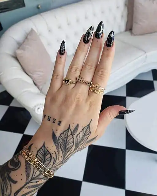 Tattooed hand with black nail art and gold rings displayed on a sofa backdrop. Elegant gold jewelry and leaf tattoo design on arm.