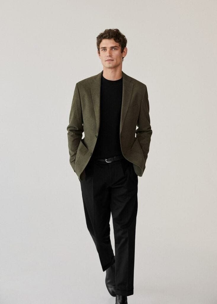 Man in a stylish olive blazer and black pants walking confidently, showcasing modern men's fashion against a plain