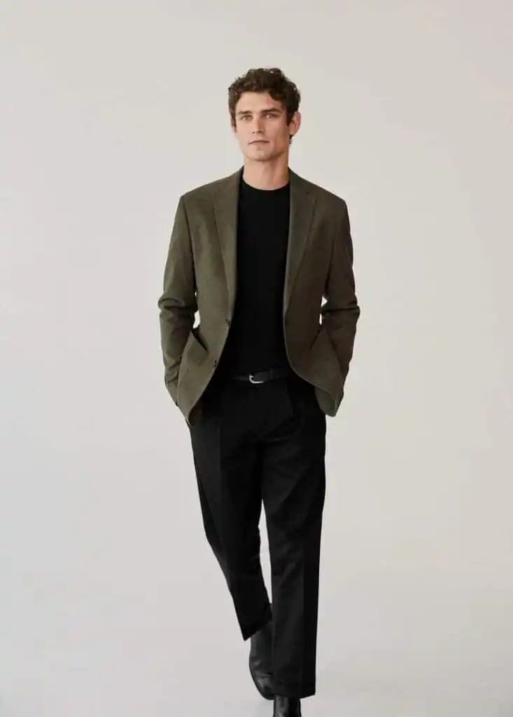 Man in a stylish olive blazer and black pants walking confidently, showcasing modern men's fashion against a plain