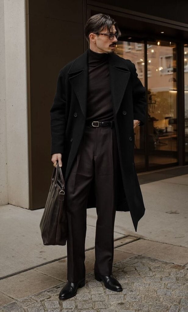 Stylish man in black coat, turtleneck, and trousers holding a leather bag, standing by a modern building entrance.
