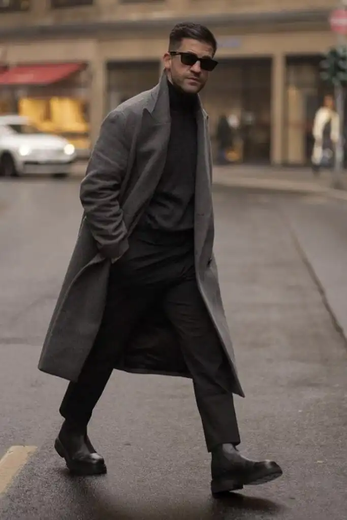 Man in stylish gray coat and sunglasses walking on city street, showcasing trendy winter fashion with confidence.