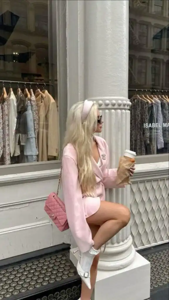 Fashionable woman in pink outfit with coffee, sitting stylishly in front of a boutique with hanging clothes.