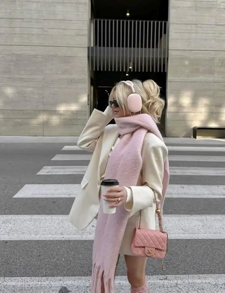 Stylish woman crossing street in pink scarf, earmuffs, and sunglasses, holding coffee and a pink purse. Urban fashion, winter style.