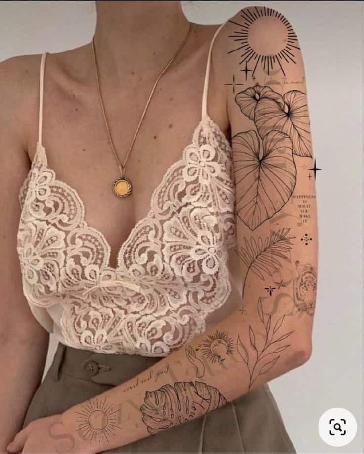 Woman in lace top with detailed floral and sun-themed arm tattoos, wearing a pendant necklace, beige trousers visible.