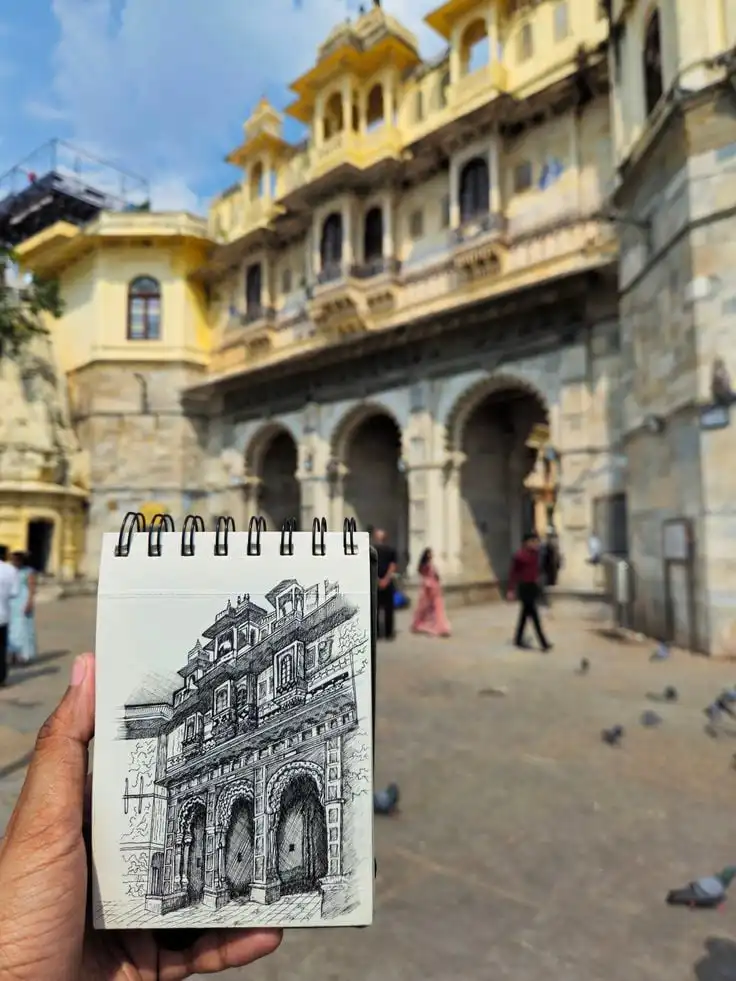 Hand holding a sketchbook with a drawing of a detailed building facade, matching the historic architecture in the background.