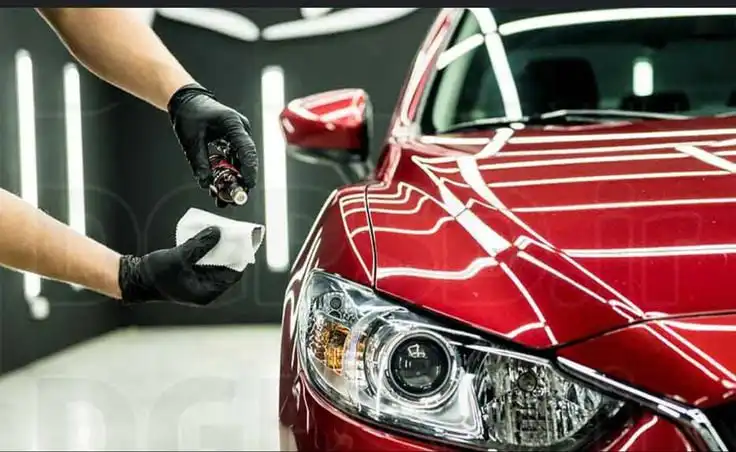 Person applying ceramic coating to red car's headlight, enhancing shine and protection in a professional detailing garage.